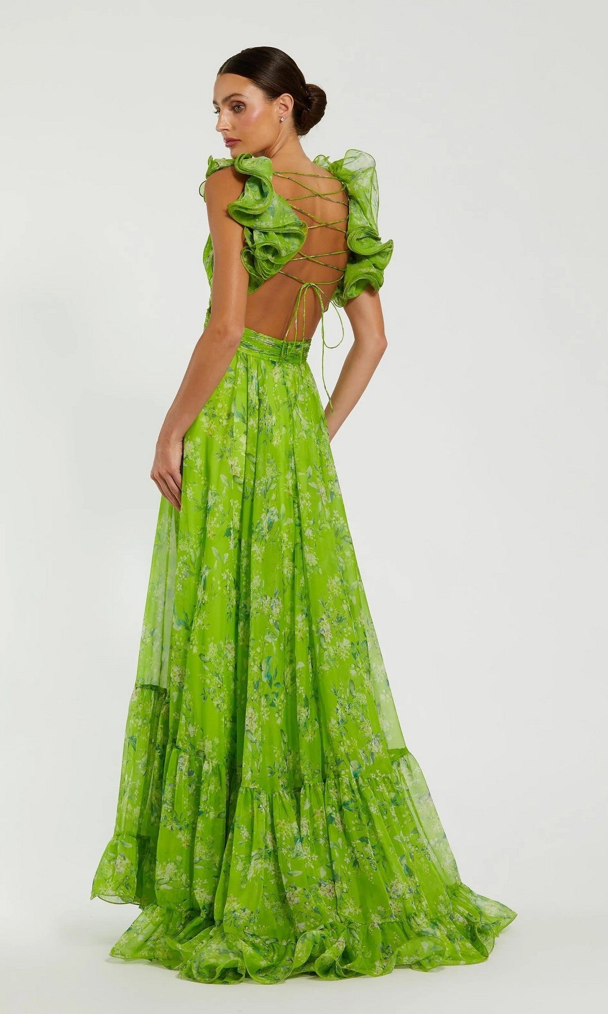 A woman stands facing away against a plain background, wearing the Mac Duggal 67803 Ruffled Long Print Formal Dress in bright green floral, with ruffled sleeves and an open lace-up back.